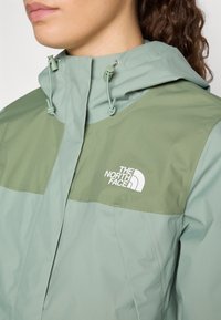 Jachetă cu glugă de culoare verde deschis, având un panou verde mai închis, cu un logo alb pe piept și șnururi la glugă. Textură netedă.