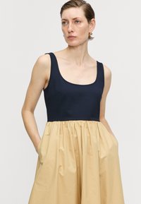 Šaty s lodičkovým výstrihom, priliehavým námorníckym živôtikom a objemnou, nariasenou béžovou sukňou. Majú bočné vrecká a hladký povrch.