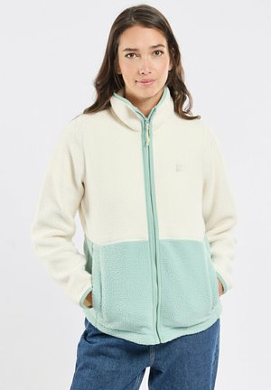 Femme portant une veste polaire zippée crème et vert clair, les mains dans les poches, debout devant un fond uni clair.