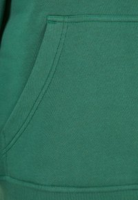 Primo piano di un tessuto di felpa verde con una texture morbida, dotato di tasca a marsupio e polsini e bordo a costine. Dettagli di cucitura visibili.