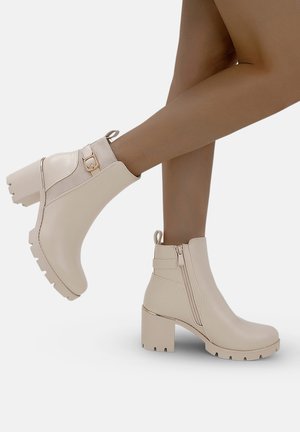 Bottines beiges avec un talon épais, en matériau synthétique brillant, fermeture éclair sur le côté et petit détail de boucle. Semelle texturée pour une meilleure adhérence.