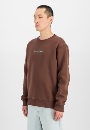 Junger Mann steht seitlich, trägt ein braunes Sweatshirt mit dem Text "Recognize the Original" und hellblaue Jeans vor einfachem Hintergrund.