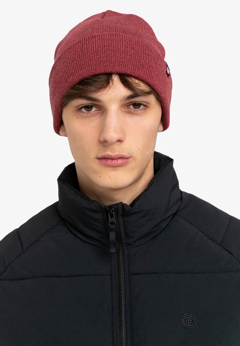 Burgundowa, dzianinowa czapka beanie z prążkowaną fakturą, z metką z logo, noszona nad czarną kurtką zapinaną na zamek z wysokim kołnierzem.