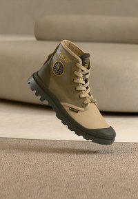 Tan and olive green high-top sneaker with rubber sole, lace-up design, logo detail on side, and contrasting toe cap. Textured fabric accents.