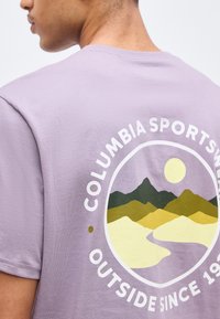 Άνδρας που φοράει λεβαντίνι μπλούζα με κυκλικό λογότυπο «Columbia Sportswear Outside Since 1938» στο πίσω μέρος, το οποίο απεικονίζει στυλιζαρισμένα βουνά, ήλιο και ποτάμι.