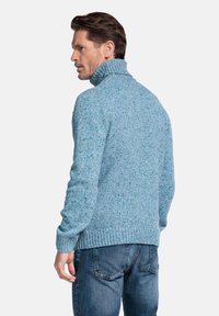 Lichtblauwe gebreide trui met een hoge kraag en geribde zoom. Het garen heeft een gespikkelde textuur. Model draagt een donkerblauwe spijkerbroek.