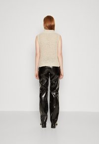 Femme aux cheveux lisses roux, portant un haut sans manches en tricot beige et un pantalon noir brillant texturé, debout face à un mur uni.