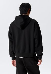 Sudadera negra con capucha, de corte holgado, con puños y dobladillo acanalados. La tela parece suave con una textura lisa.