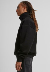 Svart zip-up hettegenser med høy krage, ribbestrikkede mansjetter og kant. Har et minimalistisk design og myk stoffstruktur. Diskret rødt logo-detalj.