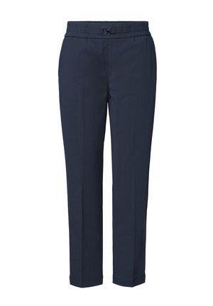 Personne portant un pantalon bleu marine fuselé, une chemise blanche et des ballerines tissées foncées, marchant vers la droite sur fond blanc.