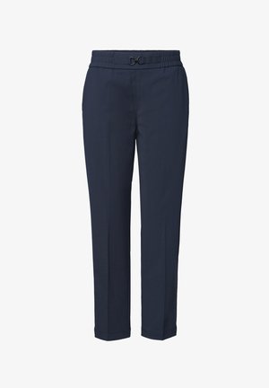 Person wearing navy blue tapered pants, white shirt, and dark woven flats walking to the right against a white background.
