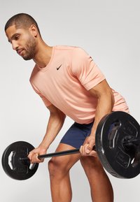 Nike lyhythihainen persikkapaita mustalla logolla, yhdistetty tummansinisiin shortseihin. Pitelee painonnostotankoa, jossa on pyöreät mustat painot, keskittynyt asento.