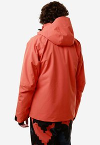 Giacca impermeabile arancione con cappuccio, dotata di una texture liscia e chiusure regolabili. Indossata con pantaloni a motivi neri e rossi.