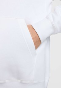 Sudadera blanca con un bolsillo frontal. Tela suave con una textura lisa. El puño presenta un detalle de costura estándar.