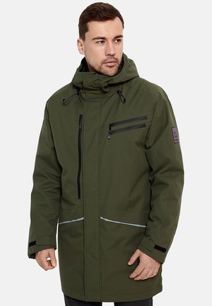 Mężczyzna w oliwkowozielonym zimowym płaszczu z kapturem, z czarnymi zamkami błyskawicznymi i odblaskowymi paskami na przednich kieszeniach, patrzący w lewo.