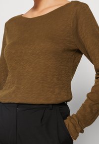 Pitkähihaine paita oliivinvihreänä, pehmeästä ja teksturoidsesta kankaasta, laajalla kaula-aukolla, yhdistettyinä mustiin housuihin. Hieno heather-kuvio.
