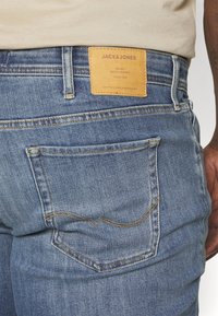 Blå denimjeans med slim fit. Har et lysbrunt lærlap med teksten "JACK & JONES" og gule søm langs baklommene.