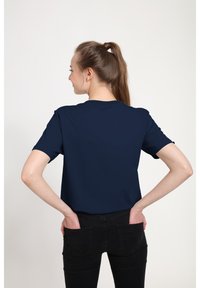 Marineblaues kurzärmliges T-Shirt aus Baumwolle mit klassischem Rundhalsausschnitt. Das Modell steht mit dem Rücken zur Kamera, die Hände in den Taschen einer schwarzen Jeans.