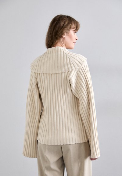 Ribbed beige knit sweater with wide collar and long sleeves, featuring vertical lines and a fitted silhouette. Worn with beige trousers.