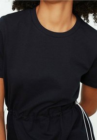 Kobieta w czarnej sukience z krótkim rękawem, okrągłym dekoltem i zaznaczoną w talii, przewiązaną białym sznurkiem.