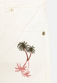 Tessuto di denim bianco con un dettaglio a tasca ornato da palme ricamate in verde e rosa. Accessori in oro vicino alla tasca.