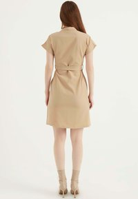 Beige Wickelkleid mit kurzen Ärmeln, einem taillierten Bund und einem knielangen Saum. Weicher Stoff mit glatter Textur und minimalen Details.