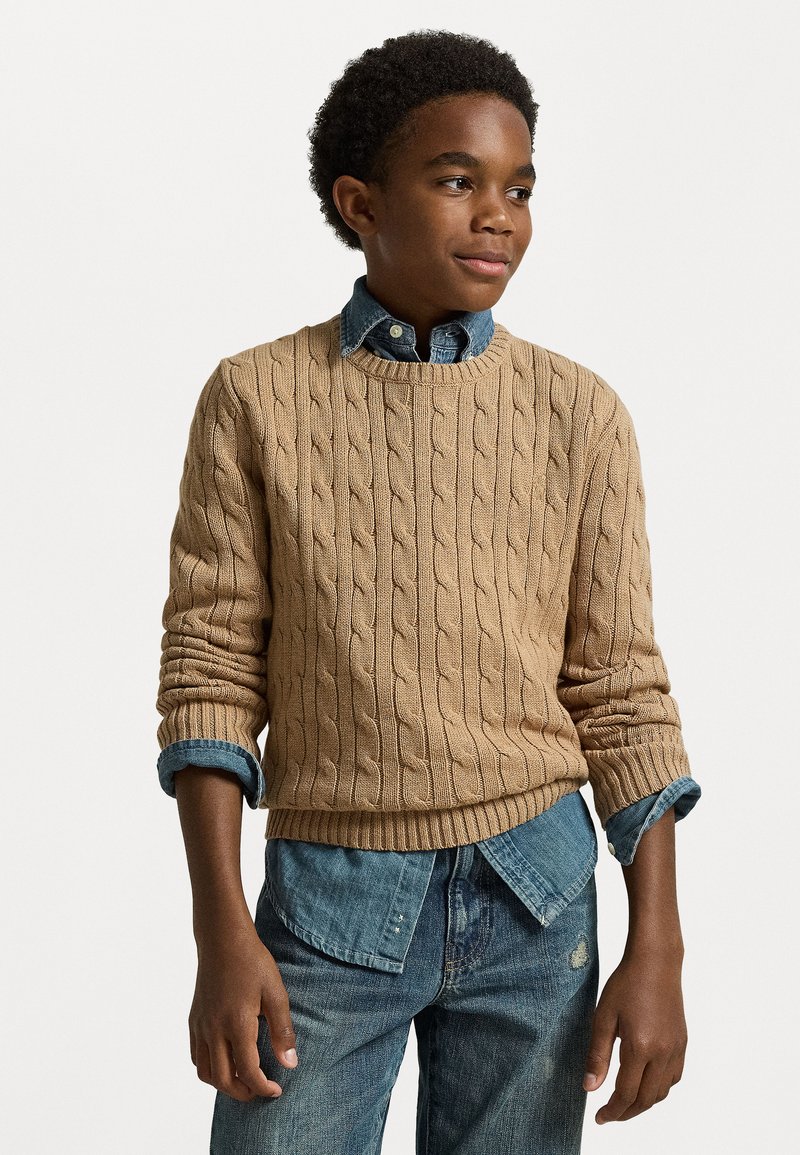 Bēšas adītas džemperis ar kabeli, savienots ar zilu denima krekli. Valkāts ar zilām džinsām, demonstrējot slāņveida ikdienišķo izskatu.