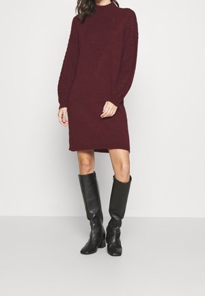 Kobieta ubrana w bordową dzianinową sukienkę z długim rękawem w połączeniu z czarnymi skórzanymi kozakami za kolano, stojąca na jednolitym białym tle.