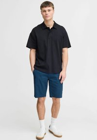 Cămașă polo neagră cu mânecă scurtă, cu guler, asortată cu pantaloni scurți teal. Modelul poartă teniși albi și șosete albe de lungime medie vizibile.
