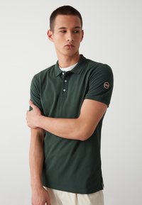 Polo de color verde oscuro hecho de una tela suave, con cuello, tres botones y un parche con el logo en la manga izquierda. Mangas cortas.