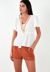 Witte blouse met korte, zwierige mouwen, voorzien van oranje bloemenborduurwerk langs de V-halslijn en een trekkoord in de taille. Gecombineerd met roestkleurige shorts.