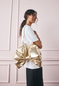 Kultainen metallinhohtoinen käsilaukku, jossa suuri rusettikuvio, sileä pinta ja vetoketjullinen suljenta. Kuvattu vaaleanpunaisella taustalla.