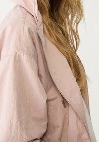 Jasnoróżowa kurtka z suwakiem z przodu, kapturem i teksturowaną tkaniną. Posiada luźny krój i widoczne przeszycia wzdłuż szwów.