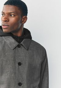 Abrigo gris texturizado con botones negros y capucha. Presenta un collar simple y una cremallera en la parte delantera bajo la capucha.