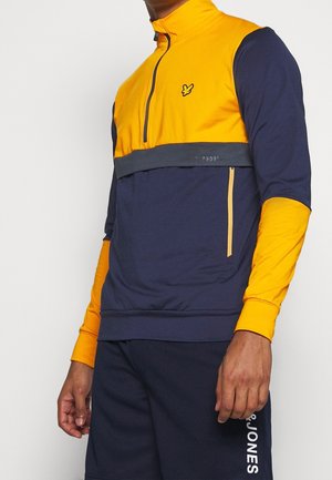 Muž nosí námořnicky modrý a žlutý pulovr na zip s malým logem na hrudi a námořnicky modré kalhoty s bílým textem na boku.