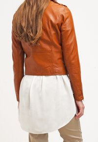 Brązowa skórzana kurtka o dopasowanym kroju, z pagonami na ramionach i gładką fakturą, zestawiona z białą tuniką.