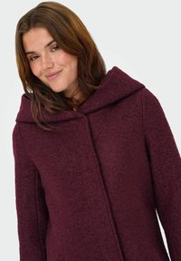 Manteau bordeaux en mélange de laine texturé. Il présente une large capuche pliée et un devant simple sans bouton. Le tissu semble chaud et confortable.