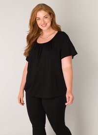 Zwarte top met korte mouwen en een ronde hals, met geplooid materiaal rond de halslijn, gecombineerd met zwarte leggings.