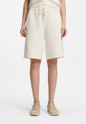 Personne portant un short crème longueur genoux avec une taille à cordon et des baskets beige clair assorties, debout devant un fond blanc uni.
