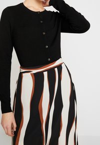 Svart stickad cardigan med lång ärm och knäppning fram, kombinerad med en randig kjol i beige, svart och brunt.
