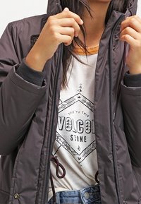 Person wearing a gray puffer jacket unzipping it to reveal a white graphic t-shirt with "Volcom Stone" text and geometric design.