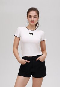 White short-sleeve top with black scalloped edges and a small black bow accent at the center, paired with high-waisted black shorts.