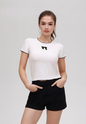 Witte top met korte mouwen en zwarte geschulpte randen, voorzien van een kleine zwarte strik in het midden, gecombineerd met hoge zwarte shorts.