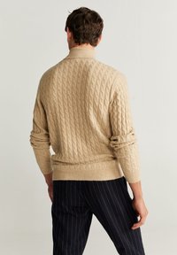 Homme vu de dos portant un pull beige en maille torsadée avec les manches retroussées et un pantalon sombre à fines rayures sur un fond uni.