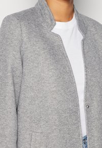 Chaqueta de punto gris con cuello mandarín, tejido texturizado y un único cierre de presión. Combinada con una camiseta blanca debajo.