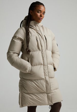 Femme aux cheveux tressés portant un long manteau matelassé beige avec des poches poitrine zippées et les mains dans les poches latérales sur un fond uni.