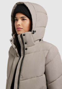 Light beige puffer jacket featuring a quilted design, zip closure, high collar, and a large hood. Black hardware accents enhance the look.
