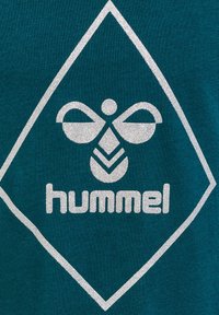 Teal stof met een zilveren logo-ontwerp met een gestileerde bij, een ruitvorm en de merknaam "hummel" eronder.