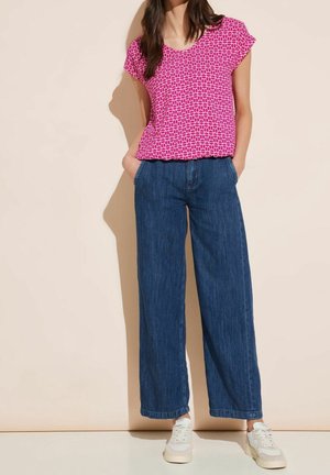 Frau trägt ein rosa gemustertes kurzärmliges Oberteil, hoch taillierte, weit geschnittene blaue Jeans und weiße Turnschuhe, steht mit den Händen in den Taschen.