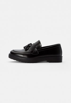Mocassin noir en cuir avec pompons, bout arrondi et semelle épaisse sur un fond gris clair uni.